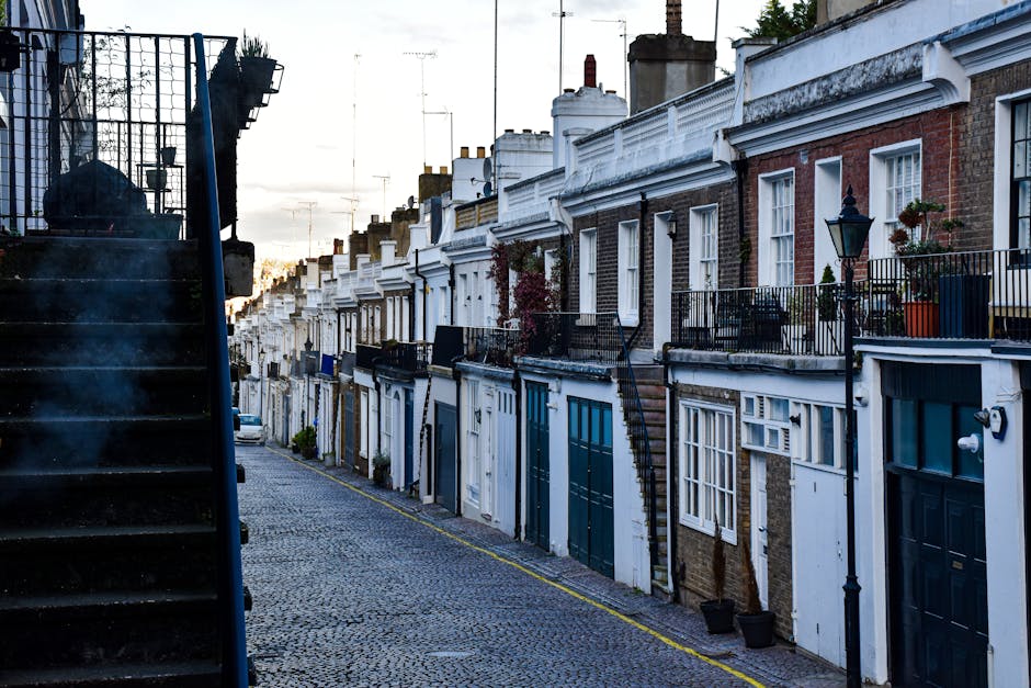 A narrow street lined with rows of terraced houses featuring brick and white-painted facades, with some houses having small balconies adorned with potted plants and decorative railings. The street is 